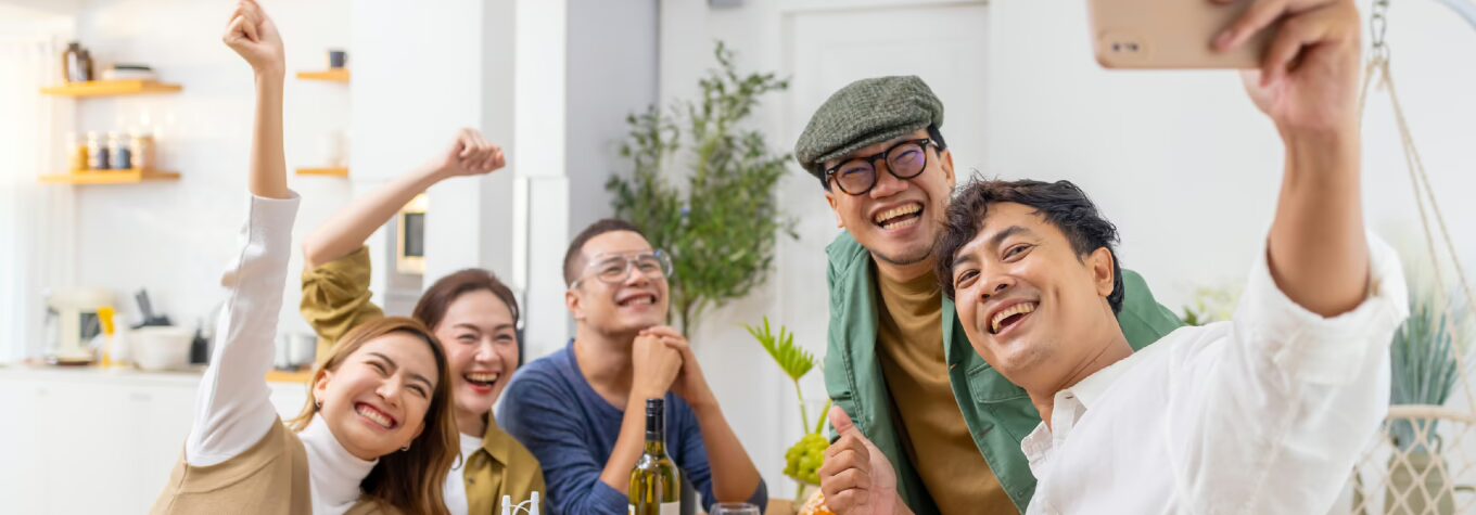 A group of five friends gathered around a dining table, smiling and posing for a selfie in a bright, modern kitchen. The table is set with food, including potatoes, vegetables, and wine, creating a warm and festive atmosphere.